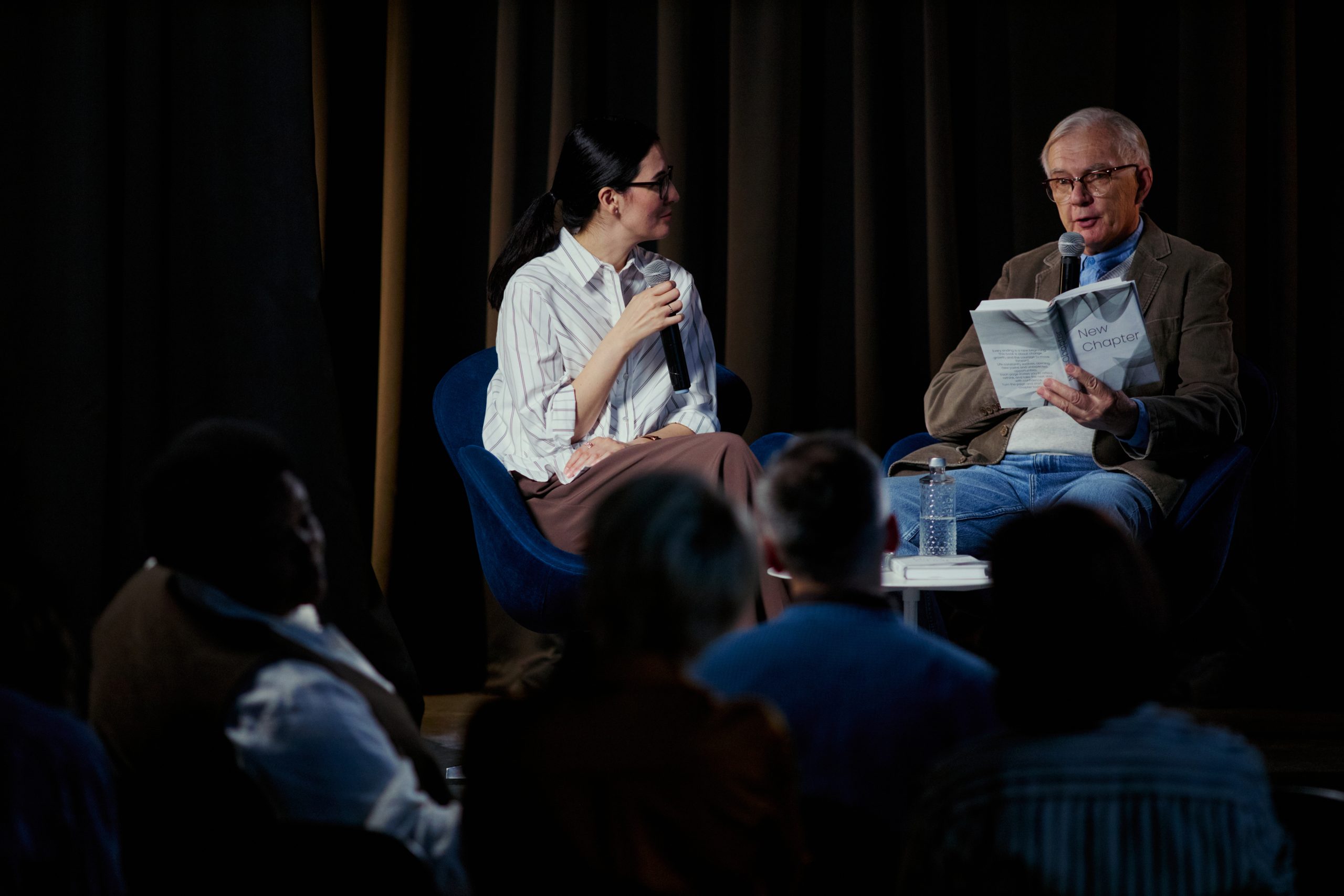 Wide shot of senior Caucasian man reading from book while young woman moderating stage discussion before audience. Ideal for publishing events, author talks, public speaking marketing